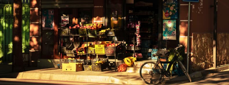 Tirana Budget Travel Local Shop and Everyday Costs Street-side fruit and vegetable shop in Tirana, Albania, with produce crates, bottled drinks, and a bicycle parked outside in the evening light.