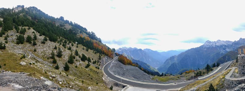 Day Trip to Theth from Shkoder Albanian Alps Road Winding mountain road leading toward Theth in the Albanian Alps, with steep rocky slopes, autumn trees, and jagged peaks surrounding the valley.