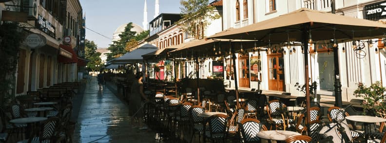 Kole Idromeno Street and Gjuhadol Shkoder Cafes Café terraces along pedestrian Rruga Kolë Idromeno and Gjuhadol in central Shkodër, Albania, with shaded outdoor tables and mosque minarets in the distance.