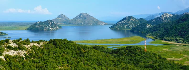Lake Shkodra Boat Tour from Shkoder Albania Views Panoramic view of Lake Shkodra near Shkodër, Albania, with calm blue water, green hills, marshy shoreline, and steep island-like peaks seen on a boat tour from Shkodër.