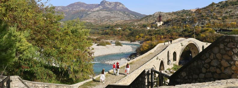 Mesi Bridge Shkoder Albania Ottoman Bridge View Mesi Bridge near Shkodër, Albania, with visitors walking across the stone Ottoman bridge above the river, framed by trees, rocky hills, and mountain scenery.