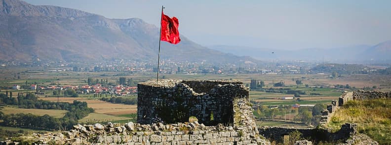 One Day in Shkoder Rozafa Castle Albania View View from Rozafa Castle in Shkoder, Albania, with the Albanian flag above the stone ruins and the city and mountains stretching across the background.