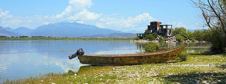 Shiroka Lake Shkodra Albania Waterfront View Wooden fishing boat on the rocky shore of Lake Shkodra in Shiroka, Albania, with calm water, lakeside buildings, trees, and mountains in the distance.