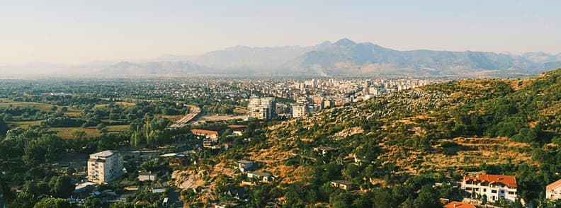 Shkoder Albania City Panorama and Mountain View Panoramic view over Shkoder, Albania, with city neighborhoods, green fields, rocky hillsides, and mountains rising beyond the urban area.