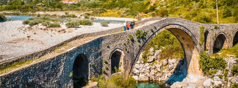 Three Days in Shkoder Mesi Bridge Albania Itinerary Mesi Bridge near Shkodër, Albania, with its high Ottoman stone arch spanning a rocky riverbed as visitors walk across the bridge.