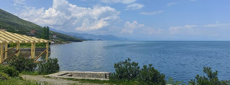 Two Days in Shkoder Lake Shkodra Albania Itinerary Lake Shkodra near Shiroka, Albania, with calm blue water, green hills, distant mountains, and a lakeside wooden structure under a bright summer sky.