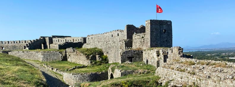 Visit Rozafa Castle Shkoder Albania Historic Fortress Stone walls and towers of Rozafa Castle in Shkodër, Albania, with the Albanian flag flying above the fortress under a clear blue sky.