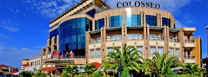 Where to Stay in Shkoder Hotel Colosseo Albania Exterior of Hotel Colosseo in central Shkodër, Albania, with its curved glass façade, palm trees, balconies, and Albanian flags beneath a blue sky.