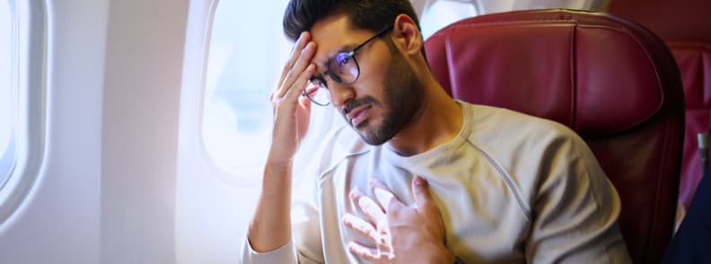 Dealing with Sleep Anxiety and Fear of Flying In Flight Relaxation Tips Man sitting in an airplane seat with a hand on his forehead and another on his chest, showing signs of flight anxiety, representing ways to manage sleep anxiety and fear of flying during travel.
