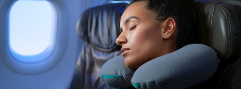 Woman resting in airplane seat with a neck pillow, illustrating how to sleep on a plane with tips for sleeping on a plane, reducing jet lag, choosing the best seat, and using accessories for comfort in economy, premium, or business class.