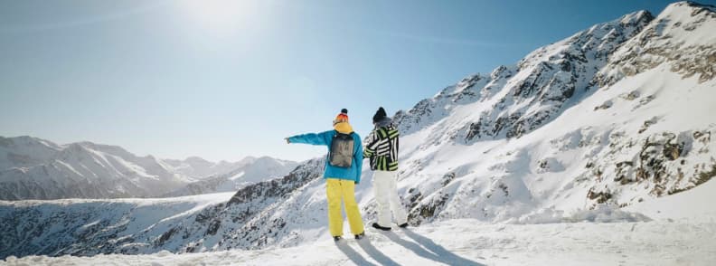 Bansko Mountain Viewpoint Bulgaria Winter Ski Trip Guide Two skiers standing on a snowy ridge in Bansko looking out over the surrounding Pirin Mountains on a sunny winter day.