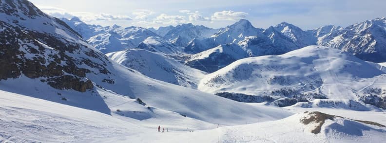 Skiing in France Alpe dHuez Grand Domaine Views Wide snowy slopes at Alpe d’Huez with skiers descending toward a backdrop of jagged Alpine peaks.