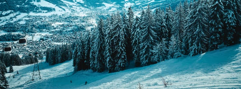 Skiing in France Megeve Forest Runs Alpine Village Views Skiers carving down a shaded forest run in Megève with the village spread across the valley below.