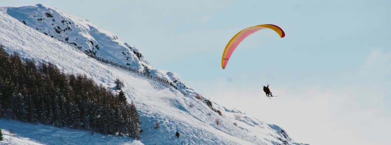Skiing in France Paragliding Les Deux Alpes Adventure Tandem paragliding flight above the snowy slopes of Les Deux Alpes with a bright wing against a clear sky.