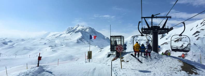 Skiing in France Tignes High Altitude Slopes Chairlift at Tignes carrying skiers toward a high altitude ridge with a French flag and wide snowy Alpine peaks.