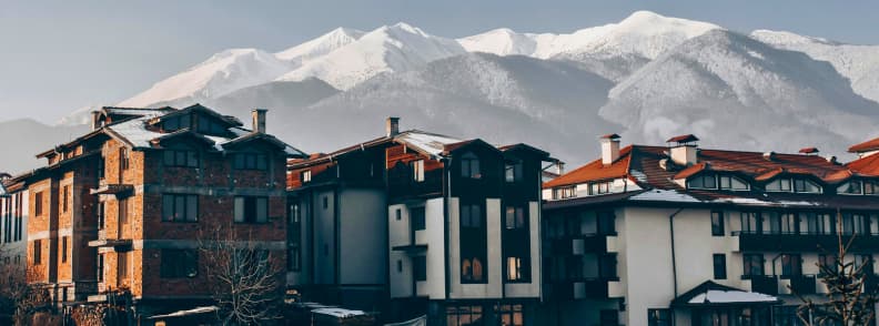 Where to Stay in Bansko Hotels Near Gondola vs Old Town Guide Hotels and apartment buildings in Bansko with the Pirin Mountains covered in snow rising behind them.