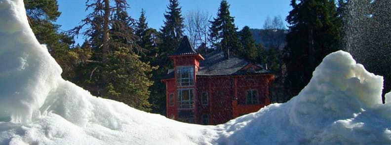 Where to Stay in Borovets Hotels and Chalets Near the Slopes Snow piled in front of a red chalet style building surrounded by tall trees in Borovets.