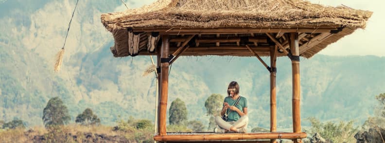 Digital nomad sitting in a bamboo hut with a camera and phone while working remotely in a tropical mountain landscape, illustrating remote work abroad and the need for flexible travel medical insurance during long-term travel.