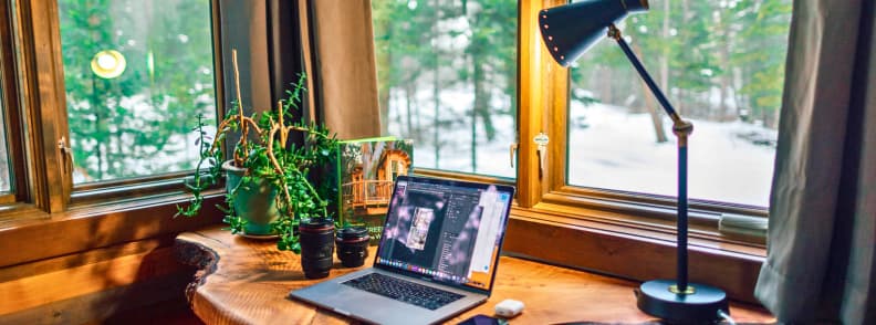 Laptop workspace on a wooden desk beside large windows overlooking a snowy forest, showing a remote work setup used by digital nomads and long-term travelers who rely on flexible travel medical insurance while working abroad.