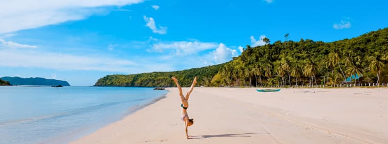 Traveler doing a handstand on a quiet tropical beach with palm trees and clear blue water, representing freedom of long-term travel and digital nomad lifestyles supported by flexible travel medical insurance.