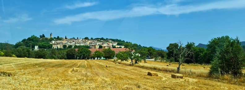 Bagnols en Foret Village and Pays de Fayence Countryside Panoramic view of Bagnols-en-Forêt hilltop village in the Var, with its church bell tower rising above stone houses and golden harvested fields dotted with hay bales.