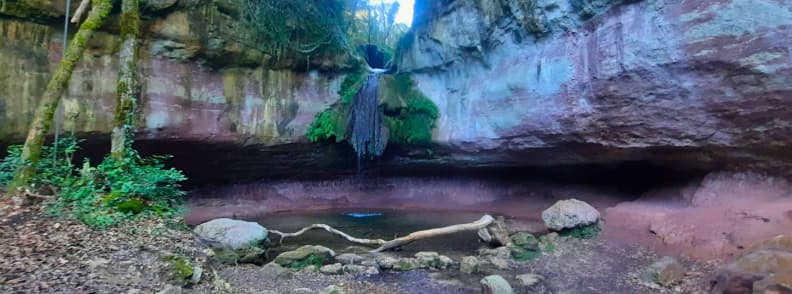 Cascade de Gourbachin Waterfall Walk Bagnols en Foret Cascade de Gourbachin near Bagnols-en-Forêt, with a thin waterfall flowing over moss-covered reddish rock into a shallow pool surrounded by stones, fallen branches, and woodland.