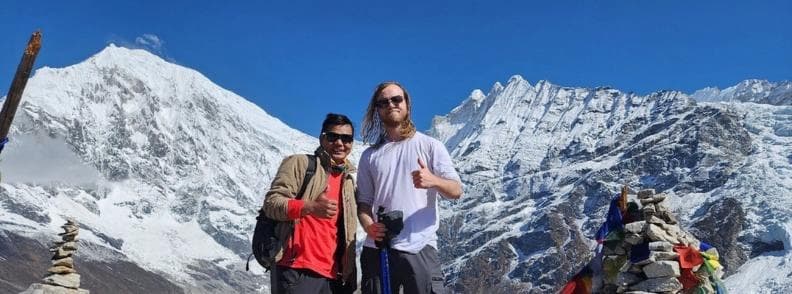 Two trekkers pose at a scenic viewpoint on the Langtang Valley Trek in Nepal, with snow-covered peaks and prayer flags in the background under a clear blue sky.
