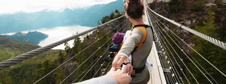 Adventure and Trust Crossing New Places While Traveling With a Baby Parent babywearing across a high bridge, hand-in-hand with partner, symbolizing confidence and connection traveling with a baby