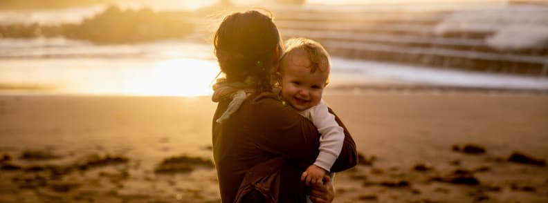 Beach Sunset With Baby Joyful Moments While Traveling with Kids Mother holding baby on beach at sunset. Smiling child enjoying golden hour during traveling with a baby on a relaxed seaside trip