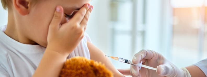 Toddler receiving vaccination before travel A toddler holding a stuffed animal receives a vaccination shot from a healthcare professional, representing health prep and vaccinations before flying with toddlers