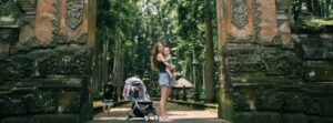 Mother traveling with a baby in Asia stands with stroller at ancient temple gate surrounded by lush forest