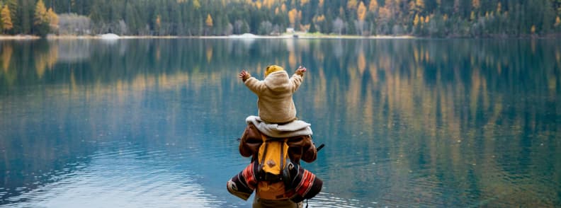 toddler traveler insurance for families Parent with toddler on their shoulders at a scenic mountain lake, highlighting the need for toddler traveler insurance during family adventures abroad