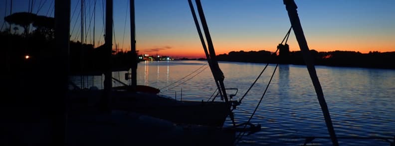 Arno River Pisa sailing lifestyle Tuscany Silhouetted sailboat rigging at dusk on the Arno River at Nautica 180 Pisa, highlighting the peaceful sailing lifestyle Tuscany offers along the upriver route to Pisa.