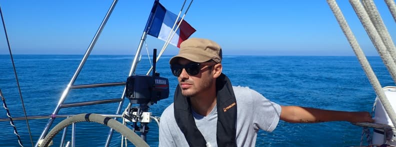 mathieu letailleur sailing holidays Italy northern Mediterranean mathieu letailleur at the helm of a sailboat during sailing holidays Italy northern Mediterranean, steering in calm sea conditions under clear skies with the French flag flying at the stern.