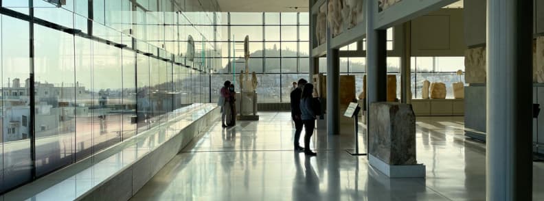 Acropolis Museum visit Athens ancient ruins Interior view of the Acropolis Museum in Athens with visitors exploring ancient Greek artifacts and panoramic views of the Acropolis hill.