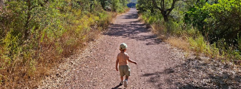 Family hike start Ravin du Gratadis Esterel Massif Saint Raphael Toddler in a sun hat walking along a wide red gravel track at the start of the Ravin du Gratadis trail from Col de Belle Barbe in the Estérel Massif near Saint-Raphaël, a family friendly route toward Col d’Aubert and Étang d’Aubert