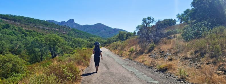 Old road return Col de Belle Barbe family hike Esterel Massif Hiker carrying a toddler in a backpack walking the old paved road toward Col de Belle Barbe in the Estérel Massif near Saint-Raphaël with hills and red slopes ahead