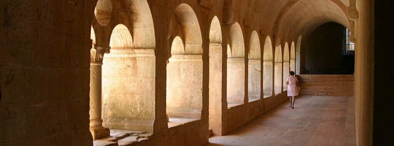 Sunlit Cloister Arches of Abbaye du Thoronet Provence Monastic Architecture Woman walking through the sunlit cloister of Abbaye du Thoronet in Provence, with a row of Romanesque arches casting shadows on the stone floor