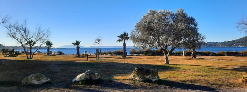 Morning view camper Etang de Berre Saint Chamas Provence View from a camper window in Saint-Chamas overlooking the Étang de Berre, with olive and palm trees in soft morning light and the lagoon glimmering in the distance.