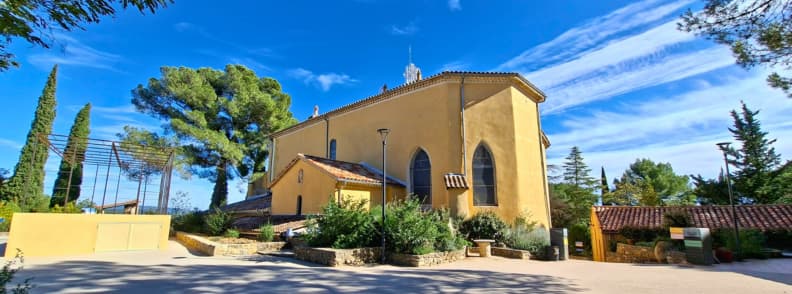 visit Sanctuary of Notre Dame de Graces Cotignac Provence Exterior view of the Sanctuary of Notre-Dame de Grâces in Cotignac Provence surrounded by cypress trees under a bright blue sky.