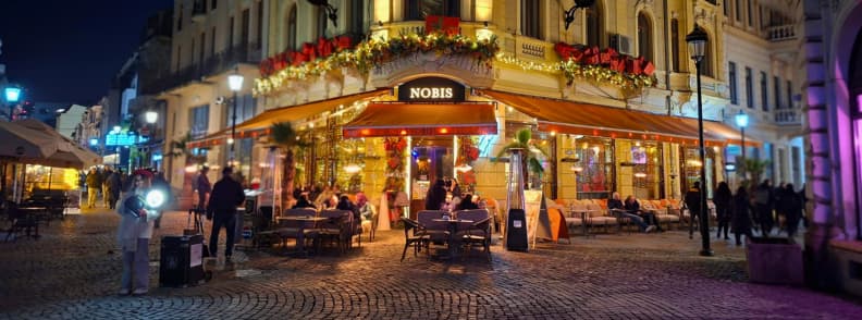 Festively decorated Nobis bistro terrace in Bucharest Old Town on a winter evening with people dining outside.