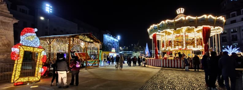 Downtown Christmas Market in Piata Universitatii with illuminated carousel, cabins, and festive decorations at night.