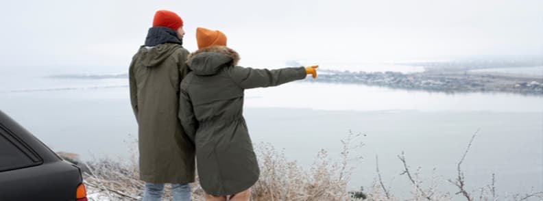 Couple standing by a car overlooking a cold European coastline during winter travel