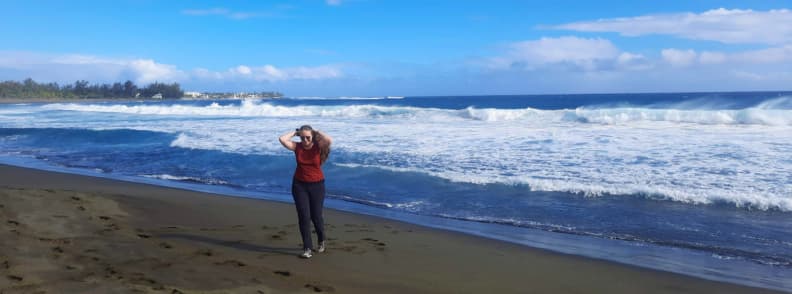 The Travel Bunny Mirela Letailleur walking on the black sand beach of Étang-Salé on Réunion Island with waves crashing behind her