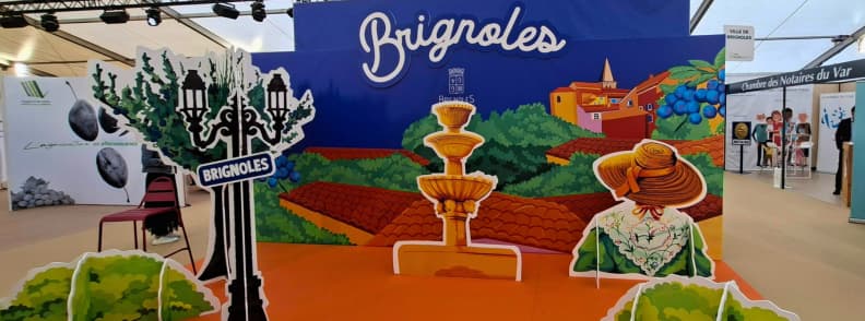 foire de brignoles 2026 dates opening hours ticket prices expo hall Decorative Brignoles-themed stand inside the Foire de Brignoles exhibition hall, with colorful Provence illustrations and exhibitor booths around.
