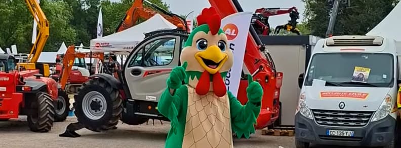 Galinou, the green rooster mascot of the Foire de Brignoles, standing among tractors and expo vehicles during the event.