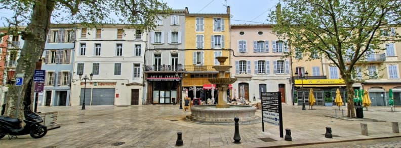 Historic square in Brignoles city center with a stone fountain, pastel buildings, and cafés around the main plaza.