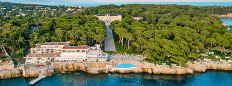 Hotel du Cap Eden Roc Near Posidonia Antibes Aerial view of Hotel du Cap Eden Roc near Posidonia Antibes, with pine forest, rocky coastline, blue sea, and the Cap d’Antibes waterfront.