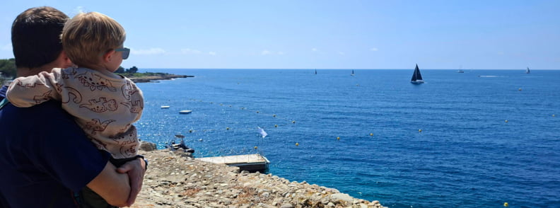 Posidonia Antibes Family Sea View Cap dAntibes Adult holding a toddler at Posidonia Antibes, looking over the blue sea, sailing boats, and rocky Cap d’Antibes coastline from a historic viewpoint.