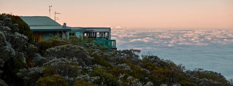 Refuge de la Caverne Dufour perched above the cloud line, surrounded by alpine vegetation at sunset near Piton des Neiges.